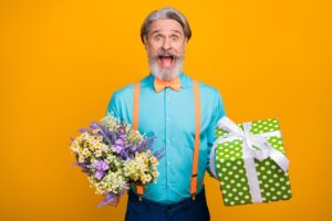 Photo of handsome grey haired grandpa hold bunch wildflowers big giftbox birthday surprise amazed wear blue shirt suspenders bow tie isolated yellow color background